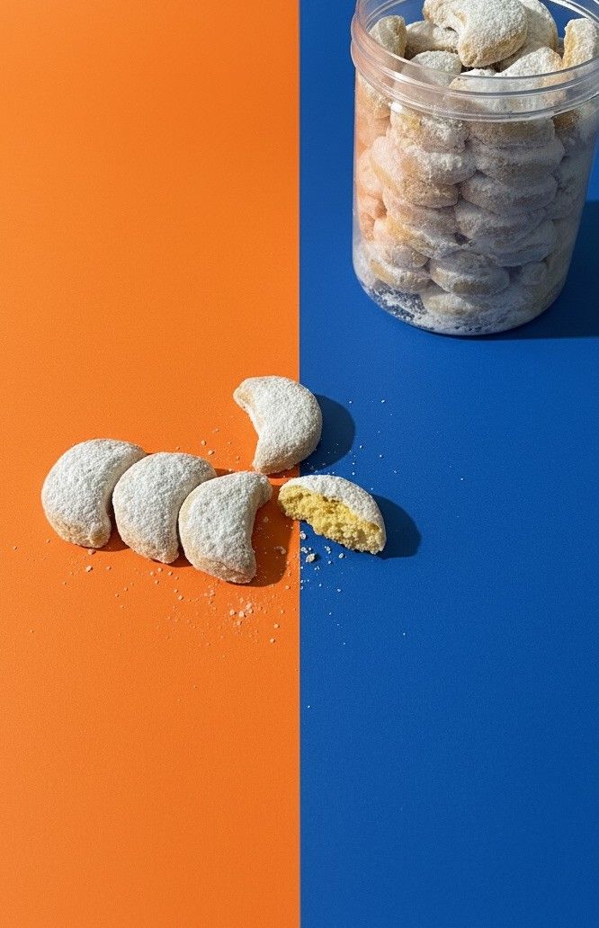Putri Salju bulan sabit bertabur gula halus, satu tergigit, di latar oranye dan biru, kue faiha.id untuk snack time di kantor